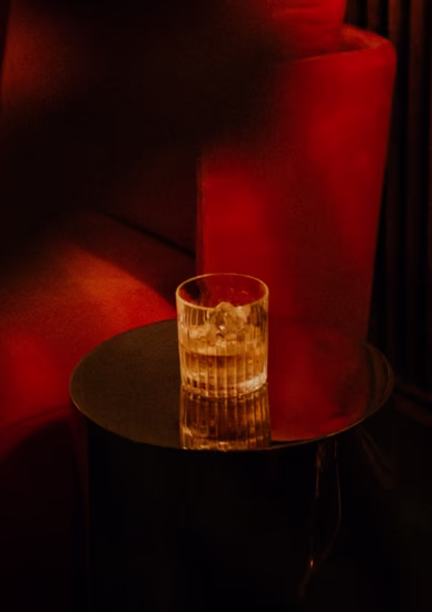Detailaufnahme eines Cocktailglases mit Eiswürfeln vor einer roten Couch in stimmungsvollem, dunklem Licht