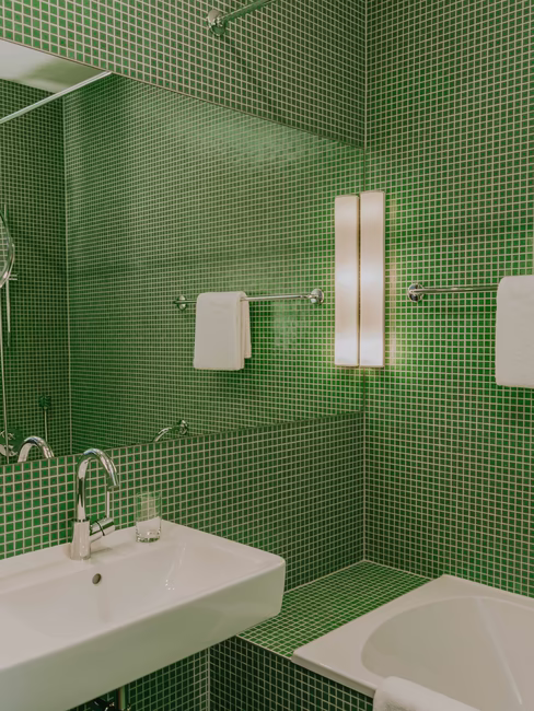 Badezimmer mit kleinen grünen Fließen und Waschbecken, Badewanne, großem Spiegel und Handtücher