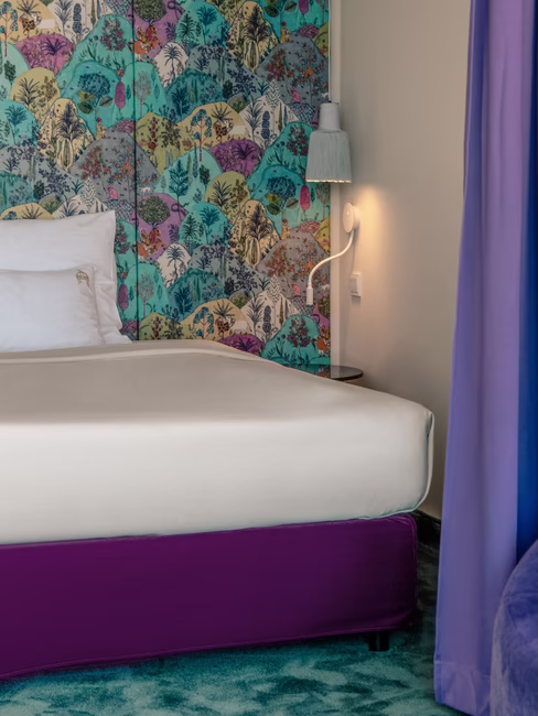 Das Bett mit farbenfrohem Hintergrund und einer davorstehenden violetten Couch.
